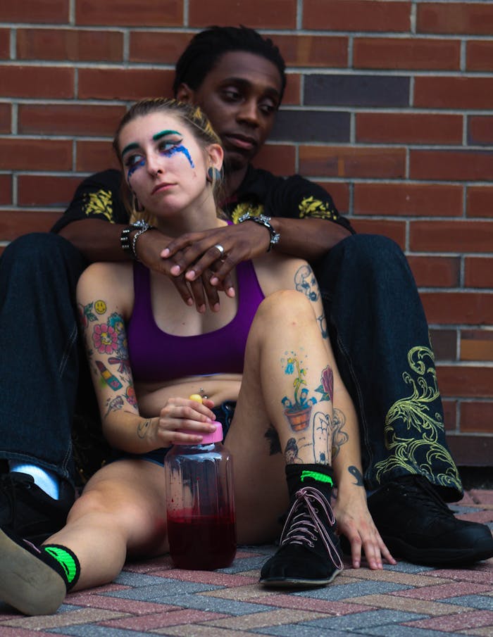 A man and woman with expressive tattoos, seated against a brick wall, depicting a deep emotional scene.