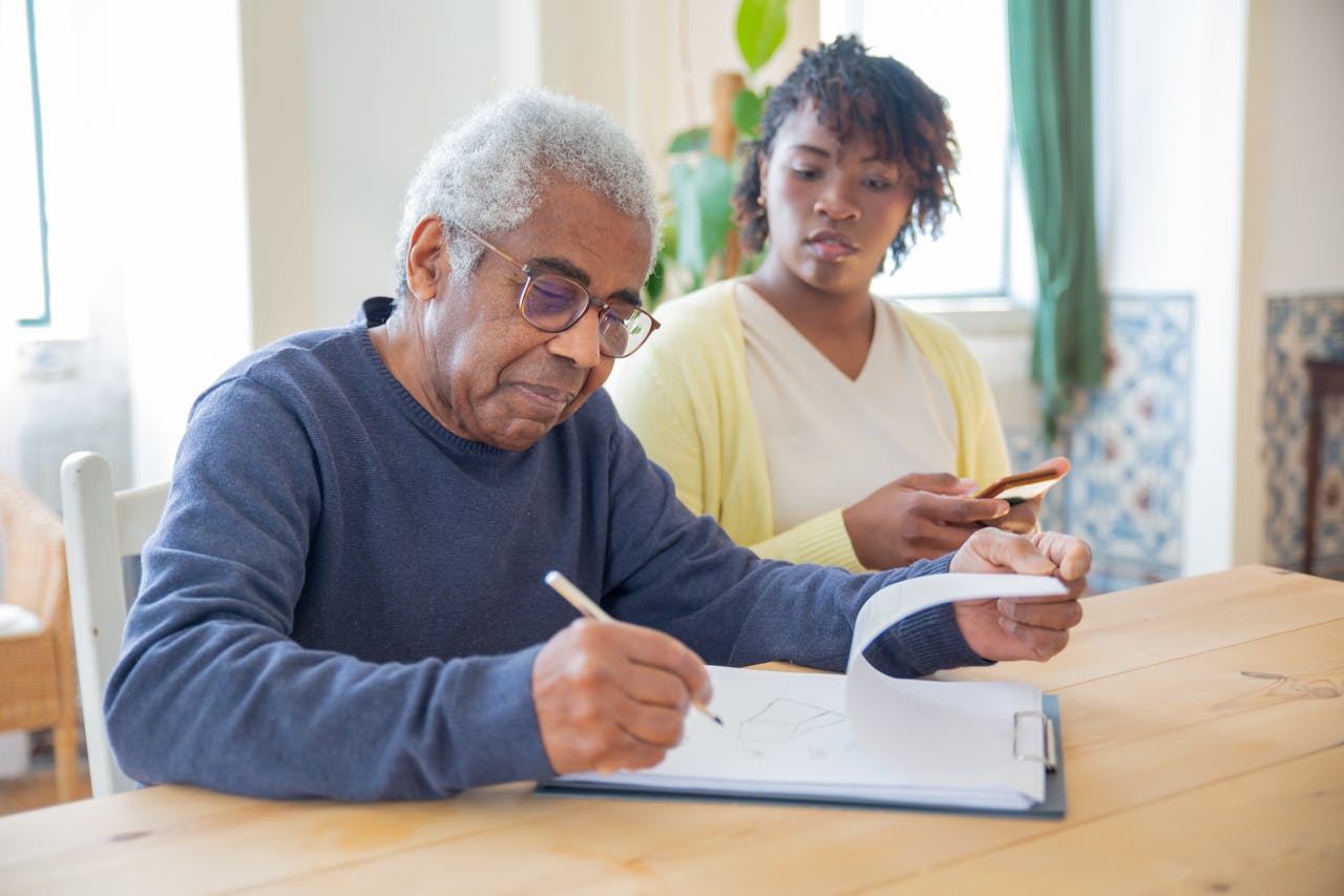 mobile-02 A senior man writing on papers with a caregiver nearby indoors in a supportive setting.